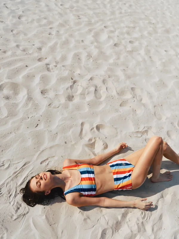 A young woman lies on bright white sand, sunbathing in a vivid striped bikini. The relaxed pose and soft shadows evoke a calm, carefree summer moment.
