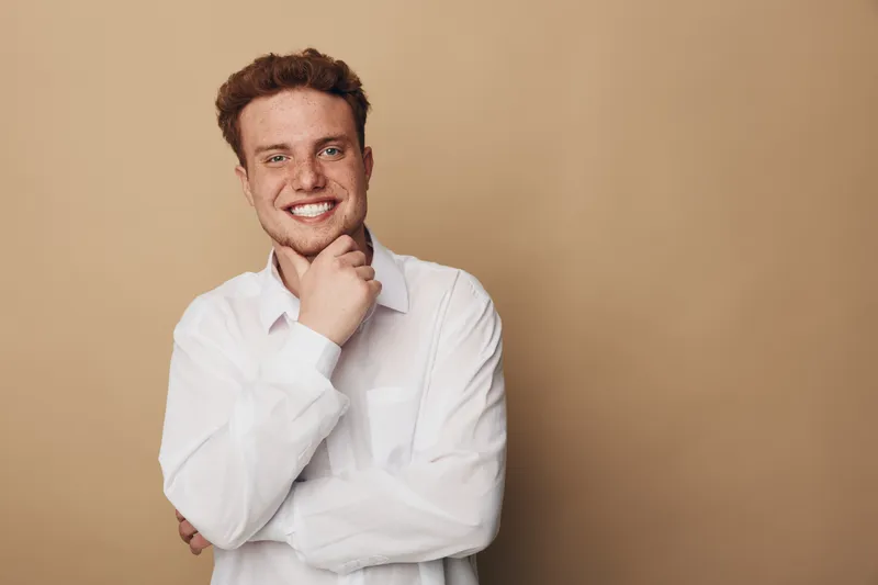 Smiling young man with red hair and freckles wearing white shirt poses with hand on chin against beige background. Concept of confidence, happiness, casual style, and positive mood.