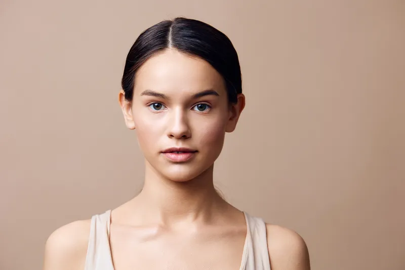 Natural beauty portrait of a young woman with a neutral expression, wearing light clothing against a beige background, ideal for wellness and beauty concepts