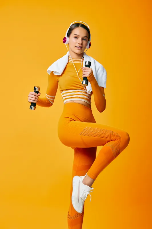 Woman fitness enthusiast in orange activewear lifts dumbbells with headphones, towel over shoulder, mid-air pose against a vibrant orange studio background, capturing energetic workout and wellness
