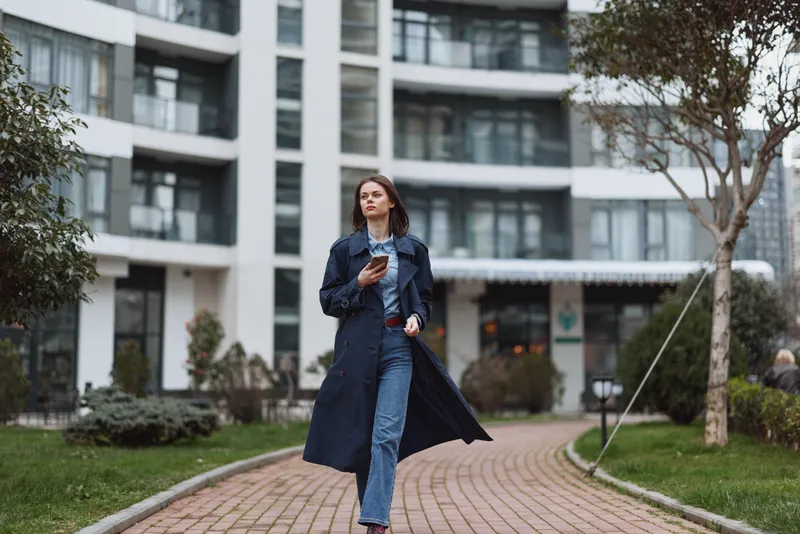 Young woman walks along a brick path in a modern city, wearing a long trench coat and jeans, holding a phone, candid urban fashion scene
