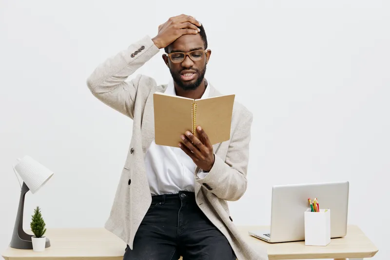 Confused and surprised young African American man in casual blazer and glasses reading a book at work station, touching his head with a puzzled expression, on plain white background, studio shot. Peop