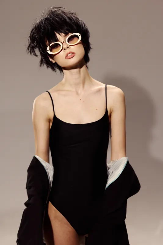 Stylish woman wearing black bodysuit and sunglasses posing with jacket on shoulders in studio lighting