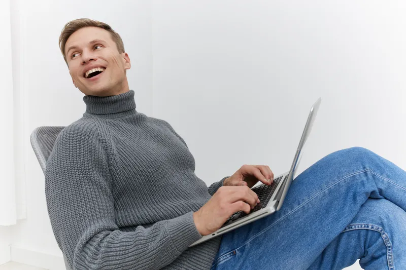 Smiling man wearing gray turtleneck sweater and blue jeans sitting on a chair using a laptop, white background, indoor studio shot, casual style, cheerful mood, technology concept.