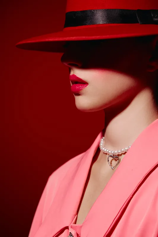 Woman with red lips wearing a pink suit and red hat hiding eyes in shadow. Pearl necklace with heart pendant. Fashion portrait with dramatic lighting on red background.