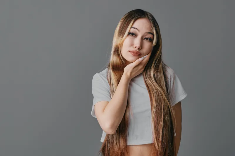 Young woman with long hair wearing a white crop top posing with hand on face against gray background for portrait or fashion.
