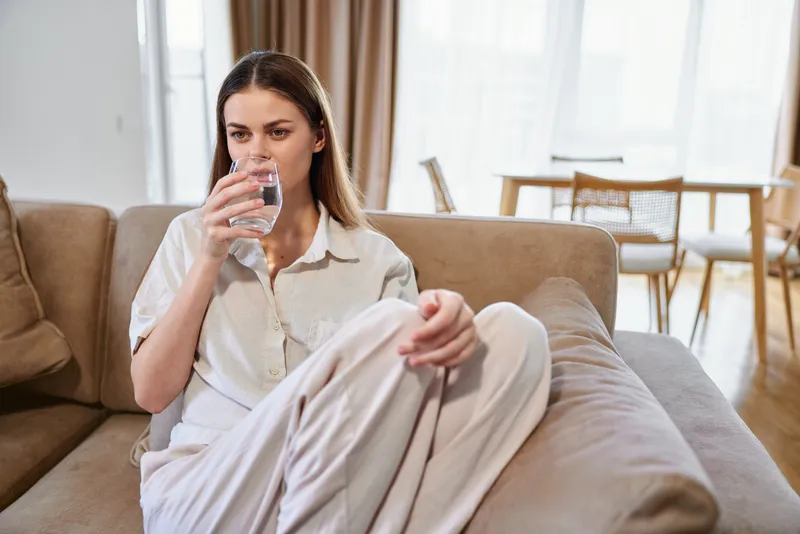 Woman drinking water from a glass on a sofa in a living room at home, relaxation moment in casual clothes with natural light, hydration, comfort and everyday domestic wellness.