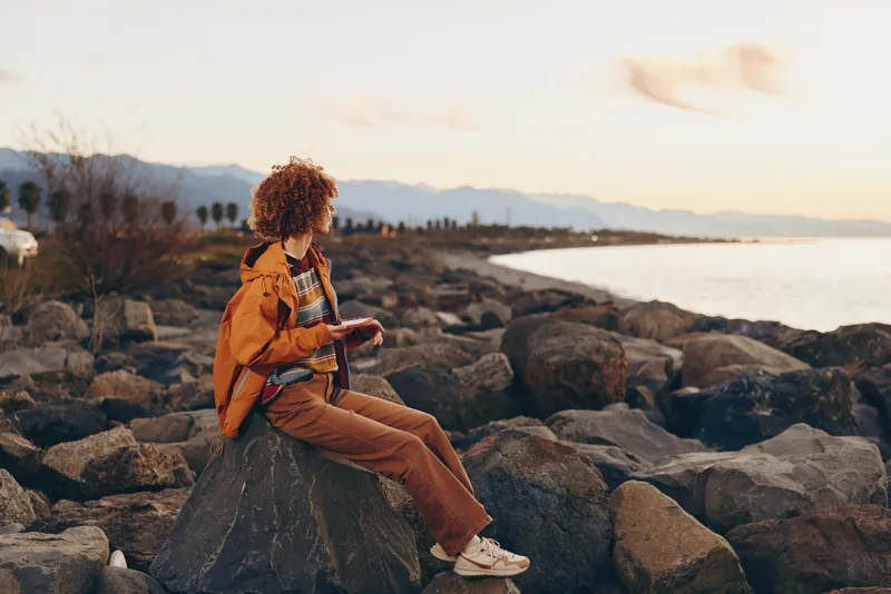 Woman lifestyle outdoors at sunset, sitting on rocky coast with backpack, curly hair moving in breeze, relaxed smile, warm golden light, ocean water and distant mountains