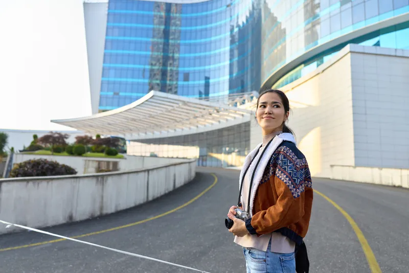 Female traveler with camera in hand stands beside modern glass buildings, urban exploration scene, casual jacket and scarf, street style photography amid futuristic architecture