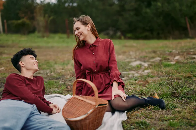 Candid outdoor picnic scene of a young couple sharing a genuine, authentic moment on a blanket with a wicker basket, laughing and relaxed in warm natural light, conveying real connection.