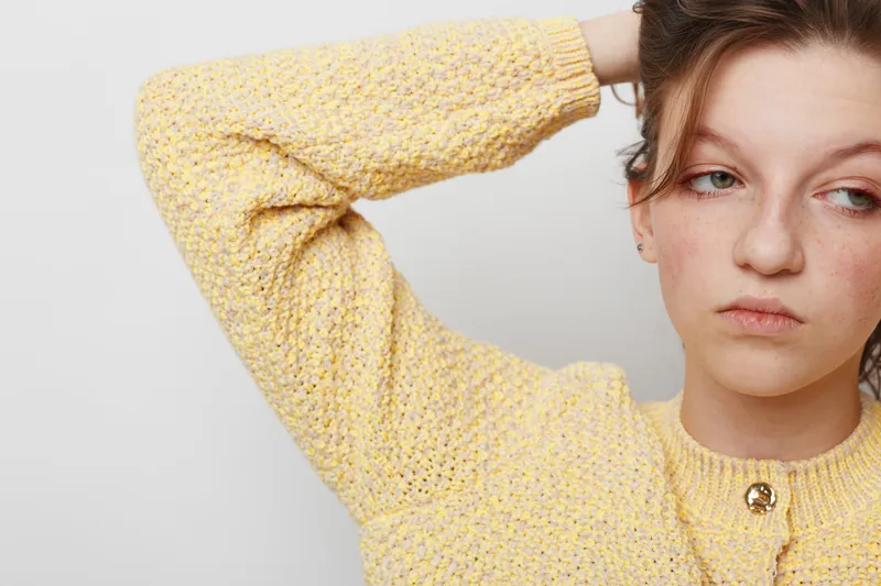 Young woman in yellow knitted sweater looks away with tired expression, hand behind head. Studio portrait on white background. Concept of fatigue, stress, and emotional exhaustion.