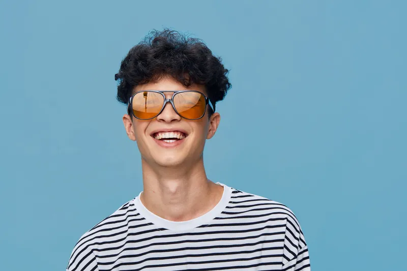 Smiling young man wearing striped shirt and stylish sunglasses stands against blue background, expressing happiness and youthful energy in casual fashion.