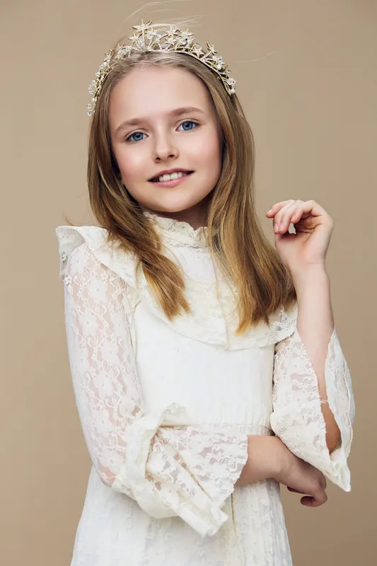 Young girl smiling with blonde hair, wearing a lace dress, and a floral crown against a beige background, exuding elegance and joy