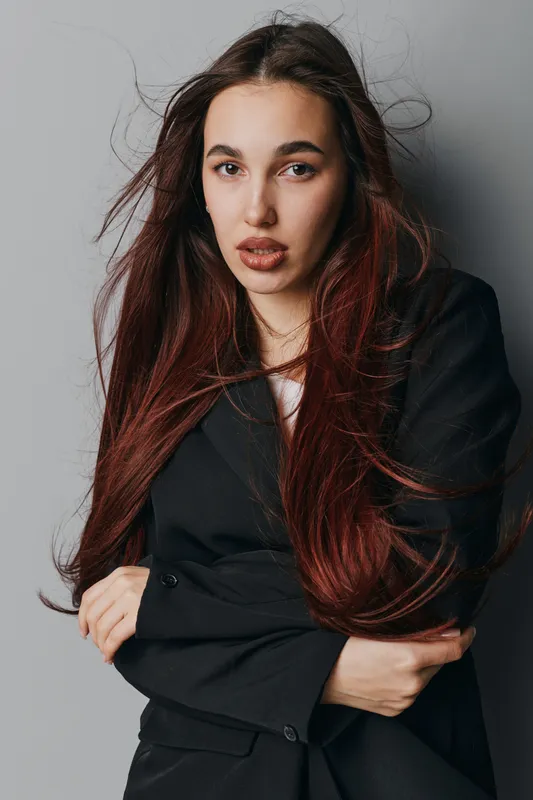 Young woman with long red hair and natural makeup posing in black blazer against gray background, looking confidently at camera with folded arms and relaxed expression.