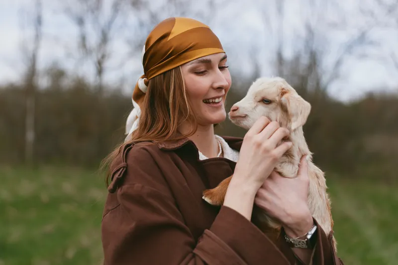 Goat kid held by smiling woman outdoors in a brown jacket and orange headscarf, affectionate countryside moment showing bonding, nature, and gentle companionship