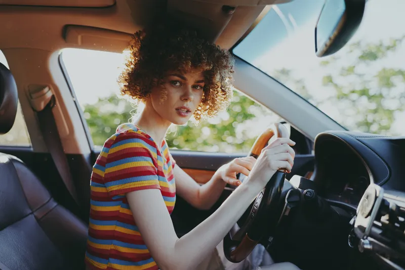 A woman with curly hair sits in the driver's seat of a car, wearing a striped shirt, looking toward the camera. Bright natural light highlights a casual, retro-inspired lifestyle.