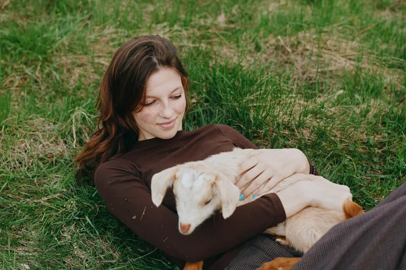 Relaxed woman lying on grass, gently cradling a young goat kid. Outdoor lifestyle moment highlighting bonding, nature, and calm companionship under soft daylight and a peaceful mood.