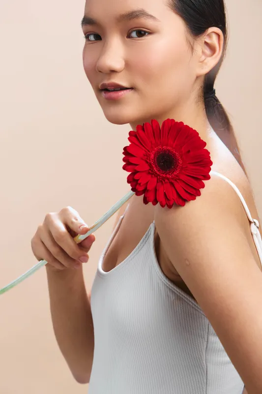 asian woman holds a bright red flower near her shoulder, wearing a white top, soft light and calm expression, creating an elegant, gentle portrait suitable for fashion and lifestyle projects.
