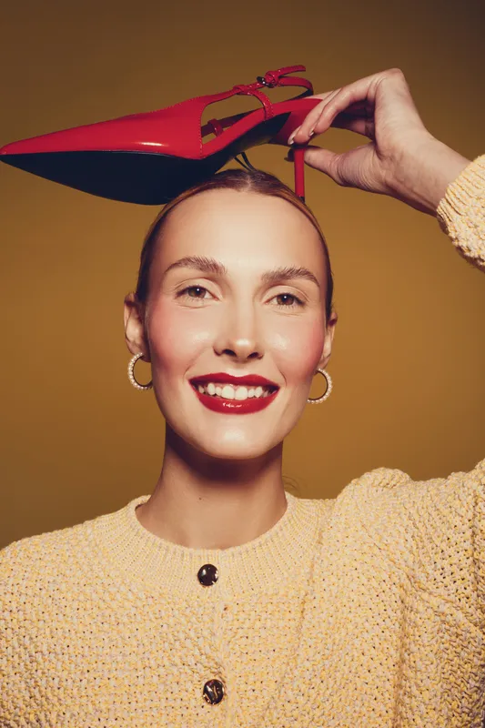 Smiling woman with red lipstick holds red high heel shoe above head. She wears yellow knitted sweater and gold hoop earrings on brown background. Concept of fashion and beauty.