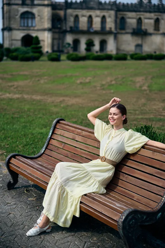 Woman in a pale yellow dress relaxing on a wooden bench in a park with a historic building in the background, creating a peaceful outdoor portrait bright day