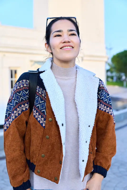 Smiling woman wearing a cozy multicolor jacket and knit sweater stands outdoors along a sunny street, showcasing casual autumn fashion and modern outerwear styling for everyday urban looks.