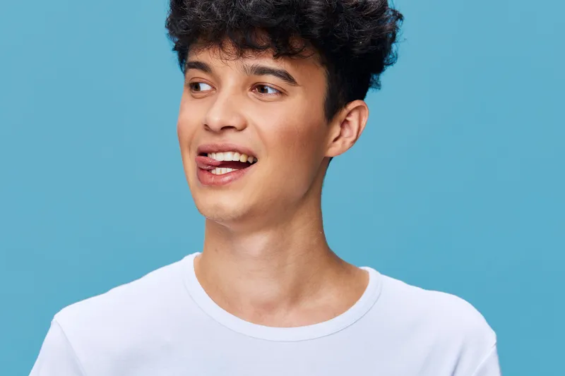 Young smiling man with curly hair wearing white t-shirt on blue background expressing joy and playfulness, close-up portrait with natural light and casual style.