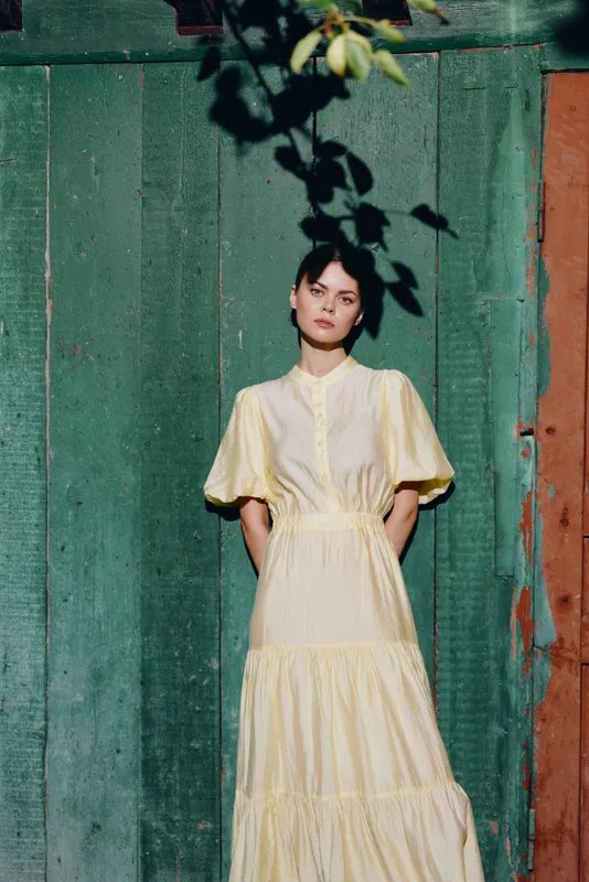 Elegant woman in a flowing pale yellow dress stands against a weathered green wooden door, posing outdoors with a calm, poised expression and soft natural lighting. An urban silhouette.