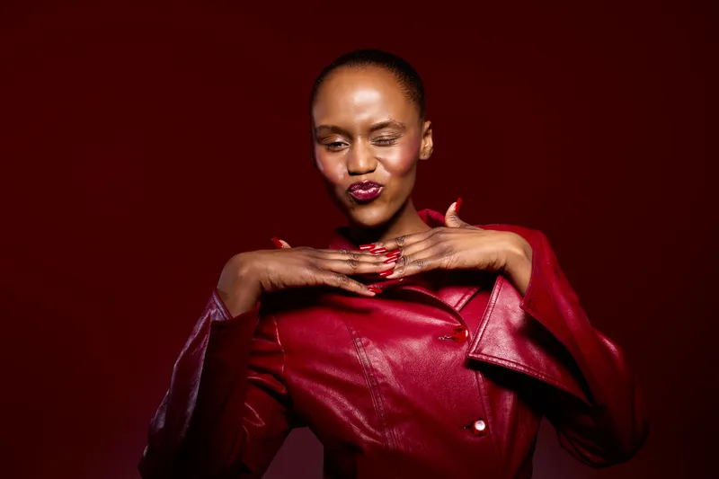 Confident African woman with dark skin poses wearing a shiny red leather jacket and matching red nails against a dark red background. Concept for fashion, beauty, and empowerment.