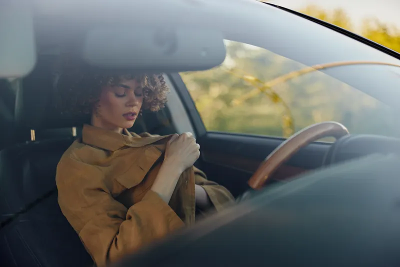 A woman inside a car wears a warm coat, adjusting it while seated in the driver's seat. Subdued retro film colors convey a calm, everyday lifestyle moment.