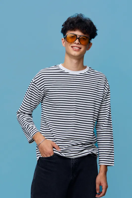 Young man wearing a striped long sleeve shirt and sunglasses posing confidently against a light blue background, smiling casually with hand in pocket.