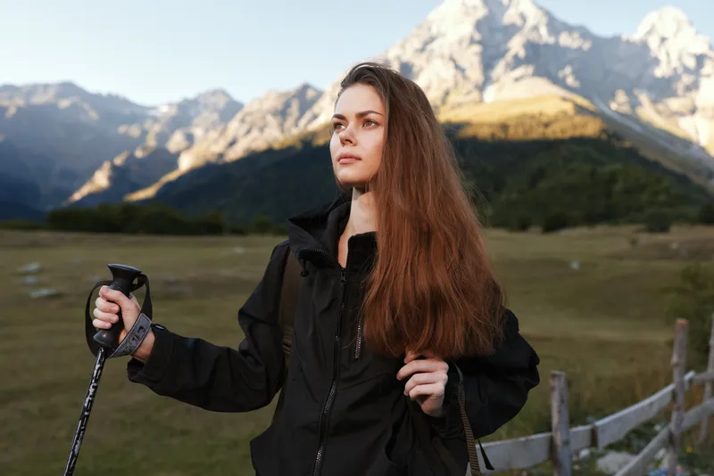 A lone female hiker stands outdoors with a trekking pole in a rugged mountain landscape. She wears a black jacket and looks ahead with calm determination, ready for adventure.
