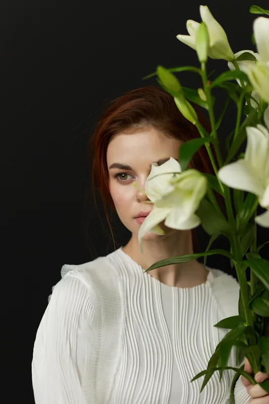 Beautiful young woman with long red hair holding a bouquet of white lilies, wearing a textured white blouse against a dark background