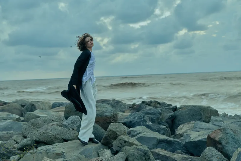 Woman stands on rocky shore by sea, wind in hair, jacket in hand, looking toward the horizon under a moody sky for travel nature and fashion inspired imagery