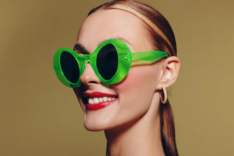 Smiling woman with green round sunglasses and red lipstick poses on beige background. Close-up portrait of stylish female model with sleek hair and hoop earrings. Fashion and beauty concept.