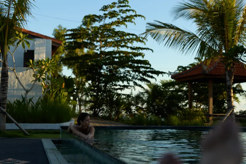 Young woman with brunette hair relaxes in a swimming pool at sunset near tropical greenery and a pavilion, enjoying a peaceful moment in a warm outdoor setting.