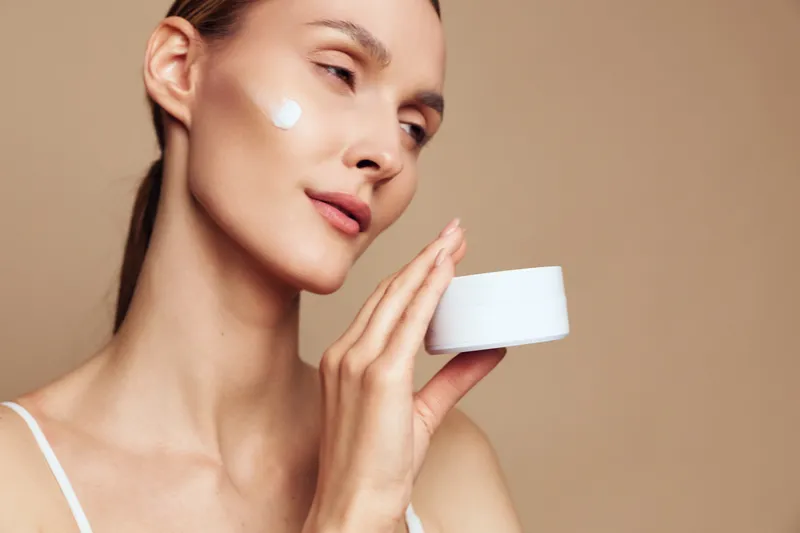 Young woman with clear skin applying white face cream on cheek, holding jar on beige background, concept of skincare, beauty routine, self-care, and moisturizing treatment.