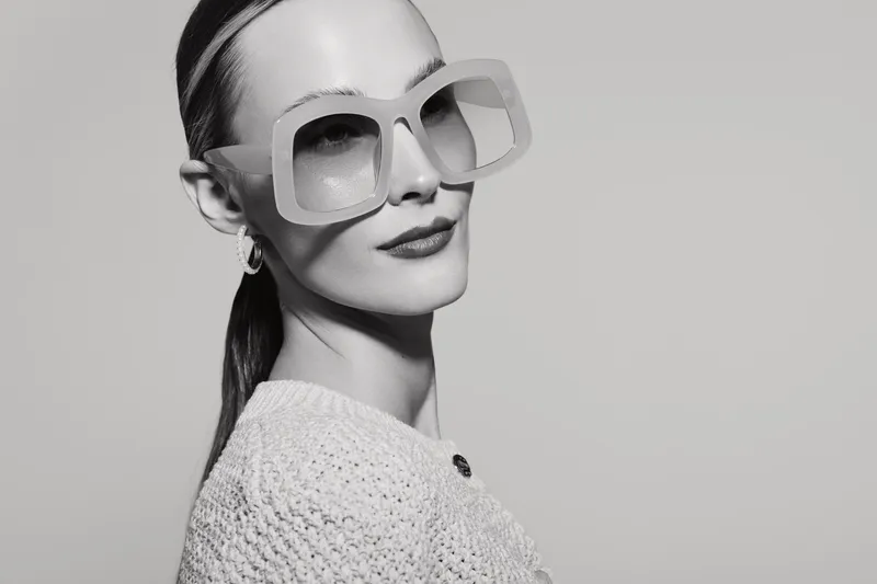 White woman with pale skin wearing large sunglasses and knitted sweater looks confidently at camera. Minimalist black and white portrait with modern fashion concept and clean background.