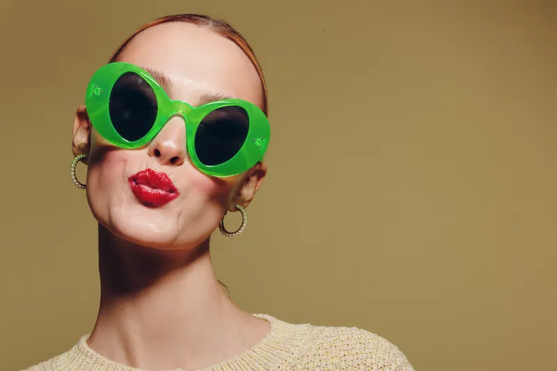 Young woman with bright red lipstick wearing large green sunglasses and hoop earrings poses with puckered lips on beige background. Concept of fashion, style, and playful attitude.