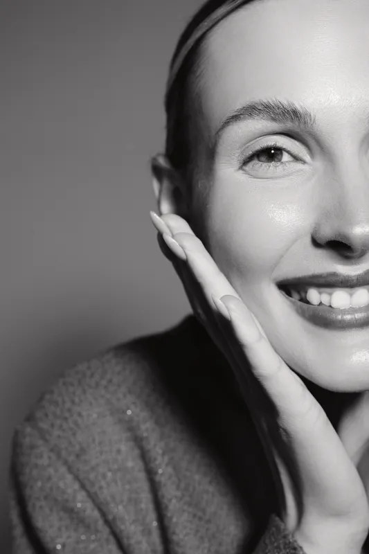 Close-up portrait of a smiling Caucasian woman with light skin touching her face, expressing happiness and natural beauty in monochrome studio lighting.