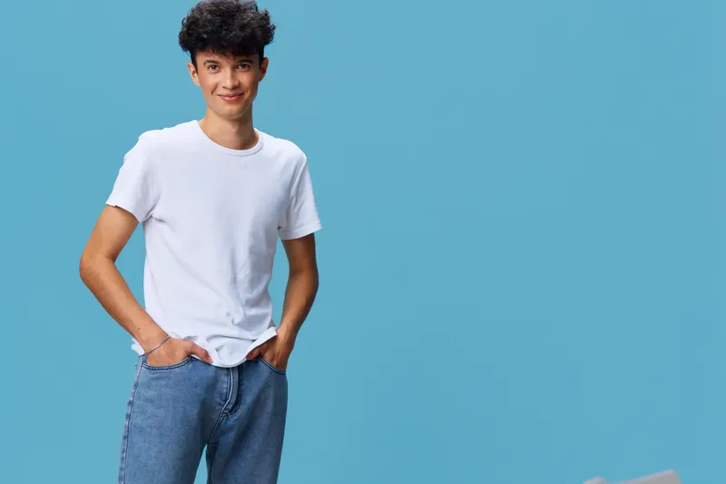 Young man in casual white t-shirt and blue jeans stands confidently with hands in pockets against a solid blue background expressing relaxed and friendly vibes.