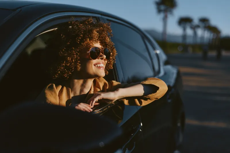 Happy woman with curly hair enjoys a sunny drive, leaning out of a car window, wearing sunglasses, in a retro film color palette, capturing a lifestyle and road trip mood.