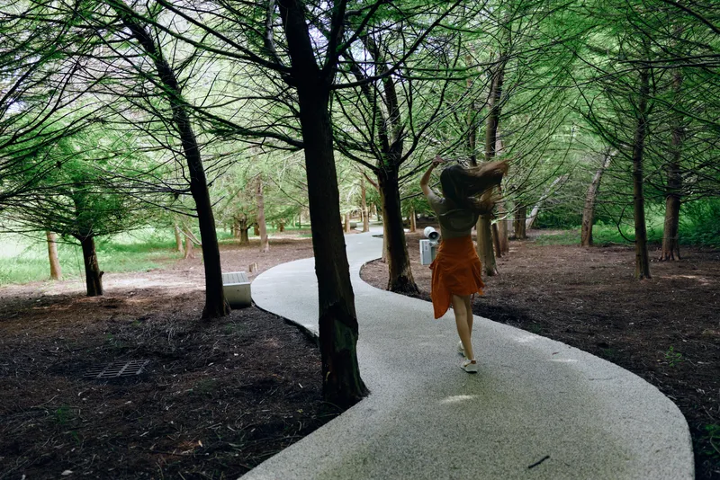 Woman walking on winding path through forest trees in a green park, wearing a sun hat and orange skirt, back view capturing motion, nature trail and peaceful summer stroll.