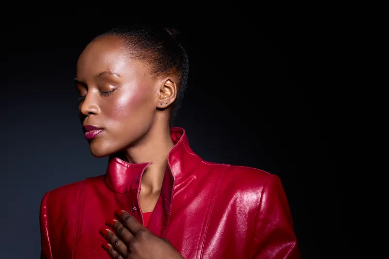 Elegant dark skinned African woman with closed eyes wears shiny red leather jacket, posing in studio with black background. Concept for fashion, beauty, confidence, and style portrait.