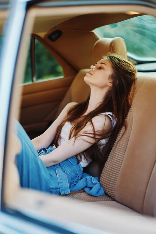 Young woman relaxing in a car, wearing a casual white top and denim jeans, exuding calm emotions in a natural green background, perfect for lifestyle and wellness concepts.
