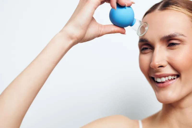 Smiling woman holding blue dropper near eye applying skincare serum, clear skin, bright background, beauty routine, natural makeup, healthy lifestyle concept