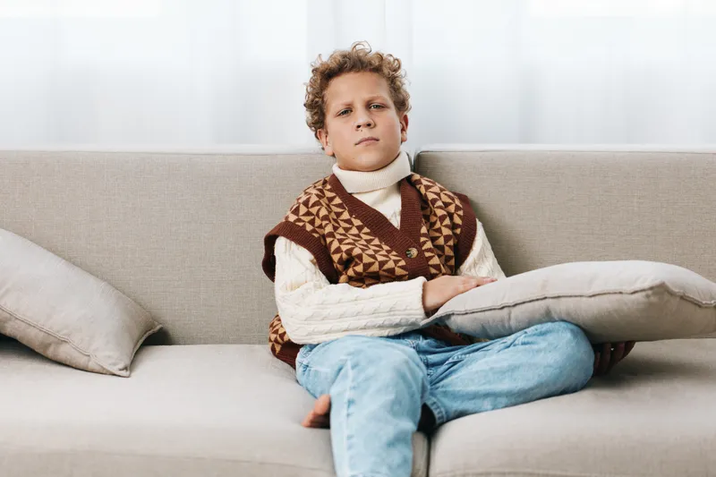 Young boy sits on a neutral sofa, wearing a knitted vest and cream sweater, relaxed pose, calm expression, warm tones, soft light, cozy indoor setting with subtle texture.