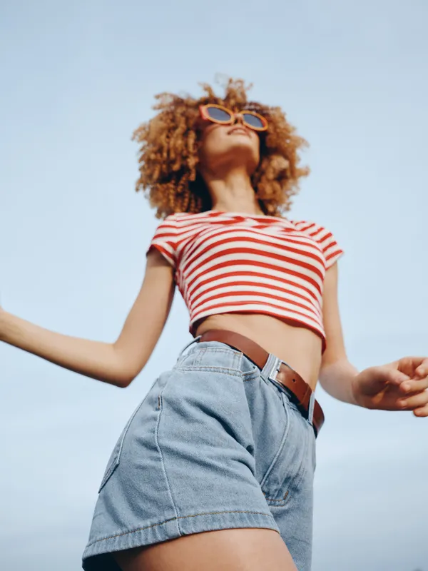 Low-angle shot of a stylish young woman in a red and white striped crop top, high-waisted denim shorts, and sunglasses, enjoying sunny outdoor fashion and freedom.