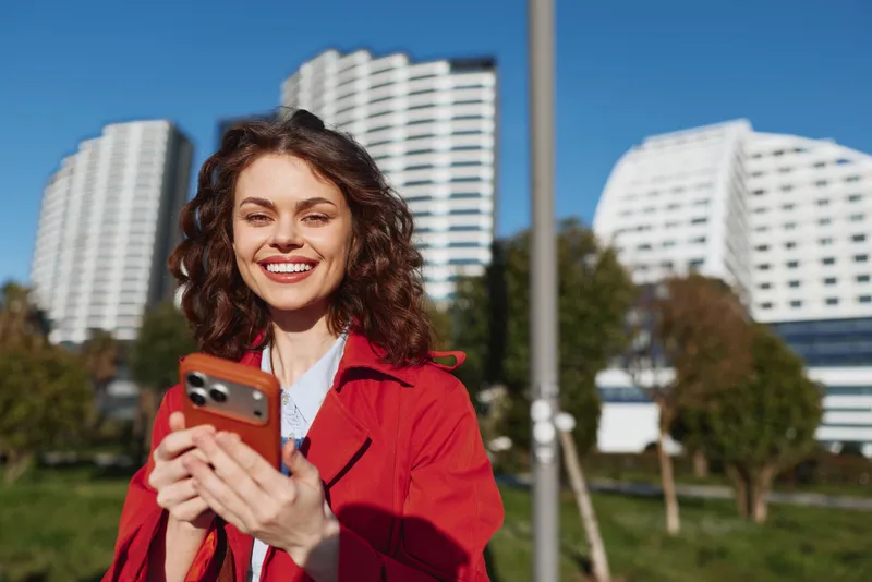 Smiling woman in a red jacket takes a selfie outdoors in a city park, with modern buildings behind, bright daytime mood, candid urban portrait.