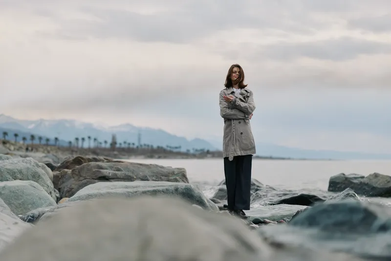 Young woman standing on rocky shore wearing cozy knitted sweater and dark pants. Cloudy sky and distant mountains create calm and contemplative mood by the sea.