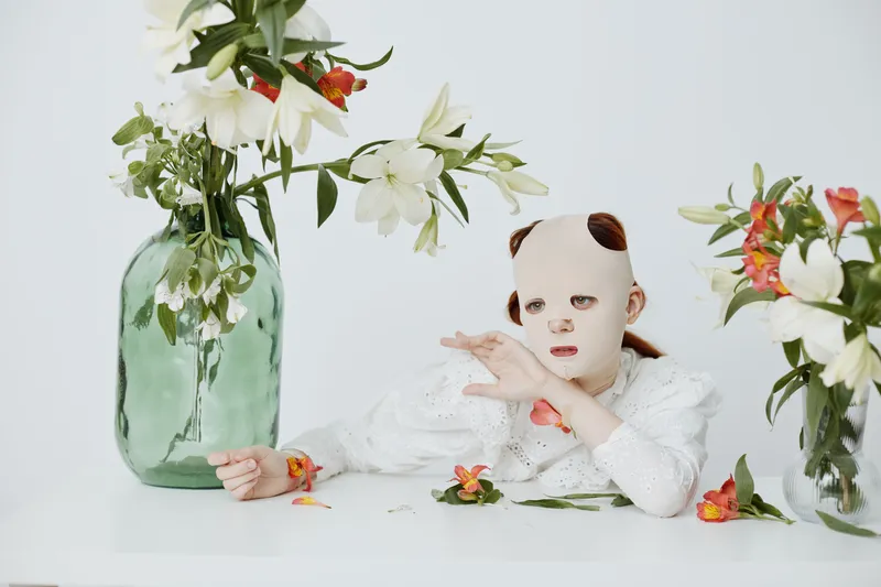 A girl with a mask surrounded by flowers, expressing curiosity in a minimalist setting with soft pastels, perfect for themes of identity and beauty